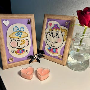 Può includere: Due pezzi di ricamo incorniciati con personaggi de La Bella e la Bestia della Disney. Il primo pezzo mostra un primo piano di Chip la tazza da tè, con un rossore rosa sulle guance. Il secondo pezzo mostra la signora Potts la teiera, con un rossore rosa sulla guancia. Entrambi i pezzi hanno uno sfondo viola e sono incorniciati in legno.