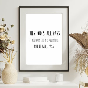 May include: A framed print with the text "THIS TOO SHALL PASS. IT MAY PASS LIKE A KIDNEY STONE BUT IT WILL PASS." The print is displayed on a white wall with a vase of flowers and a decorative stand with a gold orb on a side table.