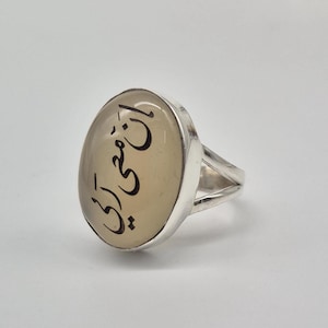Könnte beinhalten: Ovaler Ring mit einem hellen Stein und einem Silberband. Der Stein hat schwarze Kalligraphie. Der Ring hat ein geteiltes Schaftdesign. Der Hintergrund ist einfarbig weiß.