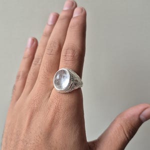 Puede incluir: Un anillo de plata con una piedra grande y transparente de forma ovalada. El anillo tiene un diseño detallado y grabado alrededor de la piedra y en la banda. El anillo se muestra en una mano.