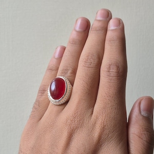 May include: A silver ring featuring a large, oval, deep red gemstone. The gemstone is set in a silver band with a textured border. The ring is on a hand, highlighting the jewellery's design and colour.
