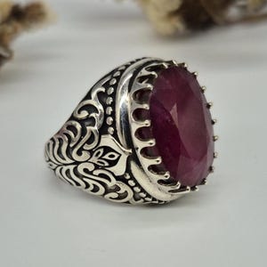 May include: A silver ring with an oval, faceted, deep red gemstone. The ring has an ornate, engraved design on the band and a crown-like setting around the stone. The ring is likely a piece of jewelry.
