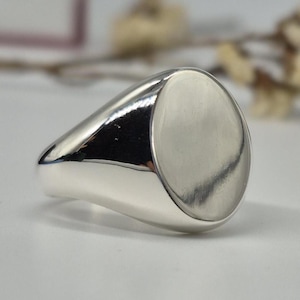 May include: A polished silver signet ring with an oval face. The ring has a smooth, reflective surface, and the design is simple and elegant. The ring is likely made of sterling silver or a similar metal. The ring is a piece of jewelry.