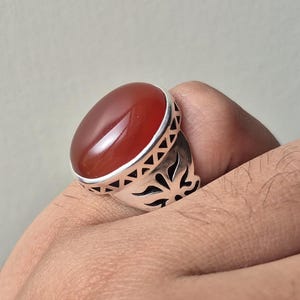 Könnte beinhalten: Ein silberner Ring mit einem ovalen, rotbraunen Stein. Der Ring hat ein dekoratives Design um den Stein und am Band. Der Ring wird von einer Hand gehalten, die das Design und die Farbe des Schmucks zeigt.