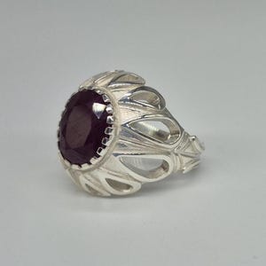 Könnte beinhalten: Ein silberner Ring mit einem großen, oval geschliffenen, tiefroten Edelstein. Die Fassung des Rings hat ein offenes, verziertes Design mit einer Reihe von geschwungenen, blattartigen Formen. Der Ring wird vor einem schlichten, hellen Hintergrund gezeigt.