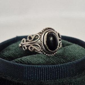 Könnte beinhalten: Ein silberner Ring mit einem ovalen schwarzen Stein, der in einer dekorativen Lünette gefasst ist. Der Ring hat verzierte Schnörkel an den Seiten und einen Perlenrand um den Stein. Der Ring wird auf einer dunkelgrünen Samtoberfläche präsentiert.