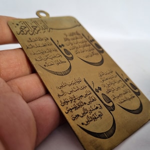 Peut inclure: Pendentif rectangulaire de couleur laiton avec des inscriptions arabes. Le pendentif comporte un petit trou en haut pour l'accrocher. Le texte est en noir et couvre toute la surface.