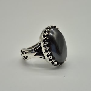 May include: A silver ring with a large, oval, dark gray stone. The ring has an ornate, openwork design on the band and around the stone. The stone is smooth and polished, reflecting light.