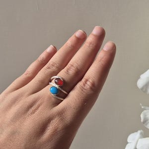 以下が含まれることがあります： 赤と青の宝石をあしらった、ダブルバンドデザインのシルバーリング。リングは手に着用され、宝石のコントラストカラーを披露しています。宝石は楕円形で、磨かれたシルバーバンドにセットされています。