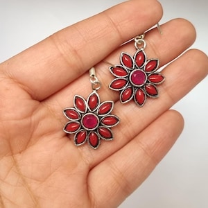 Könnte beinhalten: Ein Paar blütenförmige Ohrringe mit roten Blütenblättern und einem zentralen roten Edelstein. Die Ohrringe haben eine silberfarbene Metalleinfassung und einen Hakenverschluss. Die Ohrringe werden vor einem hellen Hintergrund präsentiert.