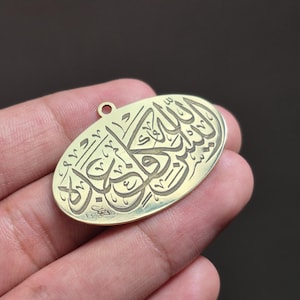 Op de afbeelding: Goudkleurige ovale hanger met Arabische kalligrafie. De hanger heeft een klein gaatje aan de bovenkant voor een ketting. De kalligrafie is ingewikkeld gedetailleerd en bedekt het hele oppervlak.