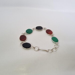 May include: A silver bracelet with oval-shaped green, red, and black gemstones. The bracelet has a clasp closure.