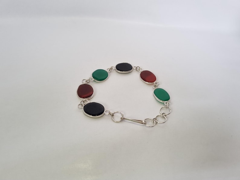 May include: A silver bracelet with oval-shaped green, red, and black gemstones. The bracelet has a clasp closure.