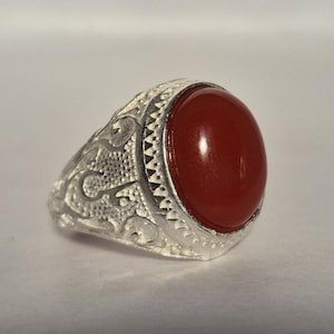 Könnte beinhalten: Ein silberner Ring mit einem ovalen, glänzenden, roten Stein. Das Band des Rings weist ein verziertes, graviertes Design auf. Der Edelstein ist der Mittelpunkt, gefasst in einer polierten Silberfassung. Der Ring wird auf weißem Grund gezeigt.