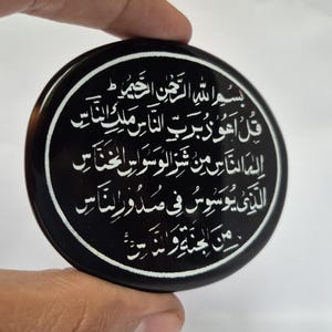 Könnte beinhalten: Ein rundes, schwarzes Dekorationsobjekt mit weißer arabischer Kalligraphie. Der Text ist kreisförmig angeordnet. Das Objekt wird von einer Hand vor weißem Hintergrund gehalten.