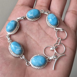 Könnte beinhalten: Ein silbernes Armband mit ovalen, hellblauen Edelsteinen. Die Edelsteine sind in einem silbernen Rahmen mit geflochtenem Rand gefasst. Das Armband wird auf einer Hand präsentiert.