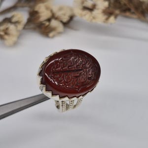 Könnte beinhalten: Ein silberner Ring mit einem ovalen, tiefroten Stein. Der Stein ist mit aufwendiger, dunkelroter Kalligraphie graviert. Der Ring wird von einem silbernen Werkzeug vor einem weißen Hintergrund gehalten. Das Band des Rings hat ein detailliertes Design.
