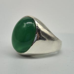 Könnte beinhalten: Ein Silberring mit einem großen, ovalen, smaragdgrünen Stein. Der Ring hat eine glatte, polierte Oberfläche und ein klassisches Design. Der grüne Stein ist der Mittelpunkt, eingebettet in ein schlichtes, elegantes Band.