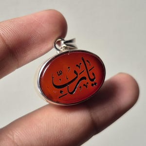Könnte beinhalten: Ovaler Anhänger mit Silberfassung und einem leuchtend orange-roten Stein. Der Stein zeigt schwarze arabische Kalligraphie. Der Anhänger wird zwischen zwei Fingern vor einem neutralen Hintergrund gehalten.