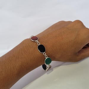 May include: A silver bracelet with four oval-shaped stones. The stones are red, black, green, and black. The bracelet is on a person's wrist.