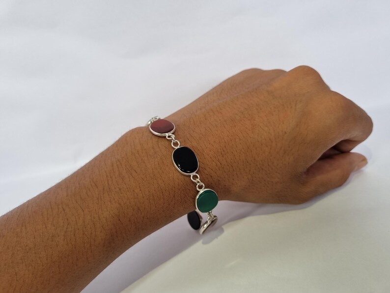 May include: A silver bracelet with four oval-shaped stones. The stones are red, black, green, and black. The bracelet is on a person's wrist.