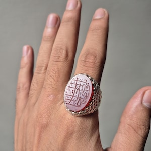 Peut inclure: Une bague en argent avec une pierre ovale translucide rose clair, montée sur une bande métallique texturée à motif de treillis. La surface de la pierre est gravée d'une calligraphie arabe complexe à l'encre foncée contrastante.