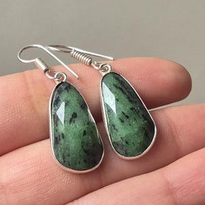 Könnte beinhalten: Ein Paar tropfenförmige Ohrringe mit facettierten grünen Steinen. Die Steine haben dunkelgrüne und schwarze Einschlüsse und sind in einem silberfarbenen Metall gefasst. Die Ohrringe haben ein einfaches Haken-Design.