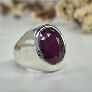 May include: A silver ring with a large, oval-cut, deep red ruby gemstone. The ruby is faceted, catching the light and displaying a rich color. The ring has a smooth, polished silver band, creating a classic and elegant look.