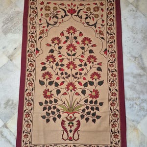 Könnte beinhalten: Eine rechteckige, beige Tapisserie mit einem bordeauxroten Rand und floralem Design. Das zentrale Motiv zeigt eine Vase mit einem Bouquet aus roten Blumen und grünen Blättern. Der Rand hat ein sich wiederholendes Blumenmuster.