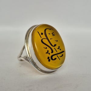 May include: Silver ring with an oval, yellow stone. The stone has black calligraphy. The ring has a silver band with an open design. The stone is set in a silver bezel.