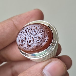 Könnte beinhalten: Ein Silberring mit einem großen, ovalen, tief rotbraunen Karneol-Edelstein. Der Stein ist kunstvoll mit detaillierter arabischer Kalligrafie und kleinen Blumenmotiven graviert. Die Ringschiene weist ein strukturiertes Kreuzschraffurmuster auf.