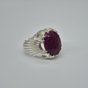 Könnte beinhalten: Ein silberner Ring mit einem großen, oval geformten, tiefroten Edelstein. Das Band des Rings hat ein strukturiertes, geschichtetes Design, und der Edelstein ist in einer dekorativen Lünette gefasst. Der Ring wird vor einem schlichten weißen Hintergrund gezeigt.