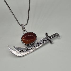 May include: A silver-toned pendant necklace featuring a large, oval, reddish-brown gemstone and a stylized sword pendant. The sword has an inscription in Arabic script. The necklace chain is a fine, textured silver.