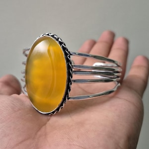 Könnte beinhalten: Ein silbernes Armreif-Armband mit einem großen, ovalen, gelben Edelstein. Der Edelstein ist in einer dekorativen silbernen Lünette gefasst und von mehreren silbernen Bändern umgeben. Das Armband wird auf einer Hand gezeigt.