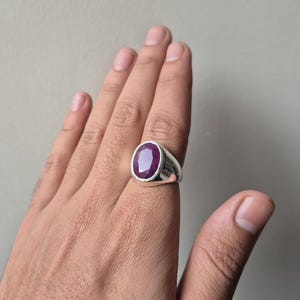 Könnte beinhalten: Ein silberner Ring mit einem großen, oval geschliffenen, tiefroten Edelstein. Der Ring hat ein schlichtes, klassisches Design mit einem polierten Silberband. Der Edelstein ist der Blickfang und fängt das Licht ein. Ein Schmuckstück.