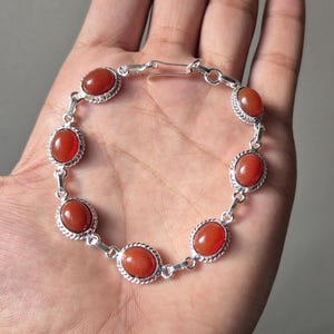Puede incluir: Una pulsera de plata con piedras preciosas ovaladas de color rojo anaranjado. Cada piedra está engastada en un bisel decorativo y texturizado, conectada por eslabones de plata. La pulsera se muestra sobre un fondo neutro.