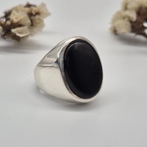 May include: A silver ring featuring a large, smooth, oval-shaped black stone set in the center. The band of the ring is polished and solid, with a wide, tapered design that supports the prominent black gemstone, creating a bold and reflective appearance.