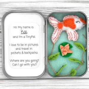 May include: A metal tin containing a felt goldfish, a small felt flower, and green felt leaves on a string. The left side features a white label with the text: Hi! My name is Kai, and I'm a TinyPal. I love to be in pictures and travel in pockets & backpacks. Where are you going? Can I go with you?