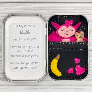 May include: A small, open tin with a felt TinyPal scene. The scene includes a pink creature and a small tan animal in a black pocket, a yellow banana, and pink hearts. The tin also has text that reads, "Hi! My name is Luna, and I'm a TinyPal."