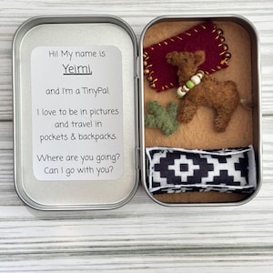May include: An open silver tin containing a felt camel, a small red felt rug, a black and white patterned fabric, and a small green felt plant. A card inside the tin reads "Hi! My name is Yeimi and I'm a TinyPal."