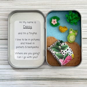 May include: An open metal tin containing a felt diorama. The diorama features a blue background with felt lily pads, a frog, and a small fabric structure. Text on the tin reads, "Hi! My name is Daisy and I'm a TinyPal."