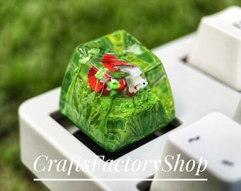 May include: A custom keycap featuring a red and white betta fish swimming in a green, resin-filled environment. The keycap is square and sits on a white keyboard. The background is blurred green. The text "CraftsFactoryShop" is at the bottom.