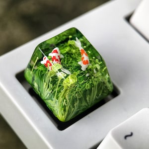 May include: A clear resin keycap with a green and white swirling design, featuring two small orange and white fish. The keycap is sitting on a white keyboard.