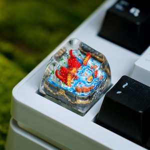May include: A clear resin artisan keycap featuring a miniature hand-painted scene of a red creature with tentacles and yellow accents set against a blue textured background, installed on a mechanical keyboard next to black and white keycaps.