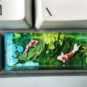 Puede incluir: Un tapón de tecla de resina con un diseño abstracto verde, azul y blanco con dos peces koi blancos y rojos nadando en el agua.