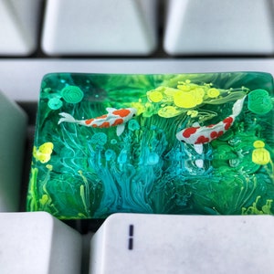 May include: A clear resin keycap with a green, blue, and yellow abstract design. Two small white and red fish are swimming in the resin.