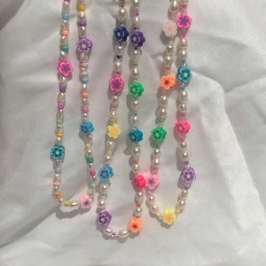 Op de afbeelding: Drie parelkettingen met kleurrijke bloemen kralen. De kettingen zijn wit met roze, blauwe, groene, gele en paarse bloemen.