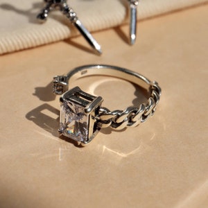 Puede incluir: Un anillo de plata con una gran piedra de circonita cúbica cuadrada, transparente, engastada en un marco de plata. La banda del anillo es una cadena gruesa de plata.