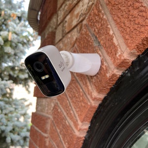 Puede incluir: Una cámara de seguridad blanca y negra montada en una pared de ladrillo. La cámara tiene una lente con un anillo negro alrededor y el texto "eufy Security" impreso en el lateral.