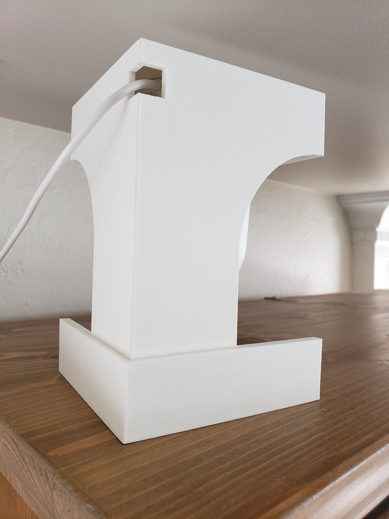 May include: A white, geometric object with an architectural design and curved features. A white cable emerges from a square aperture at the top. The object rests on a wooden surface.